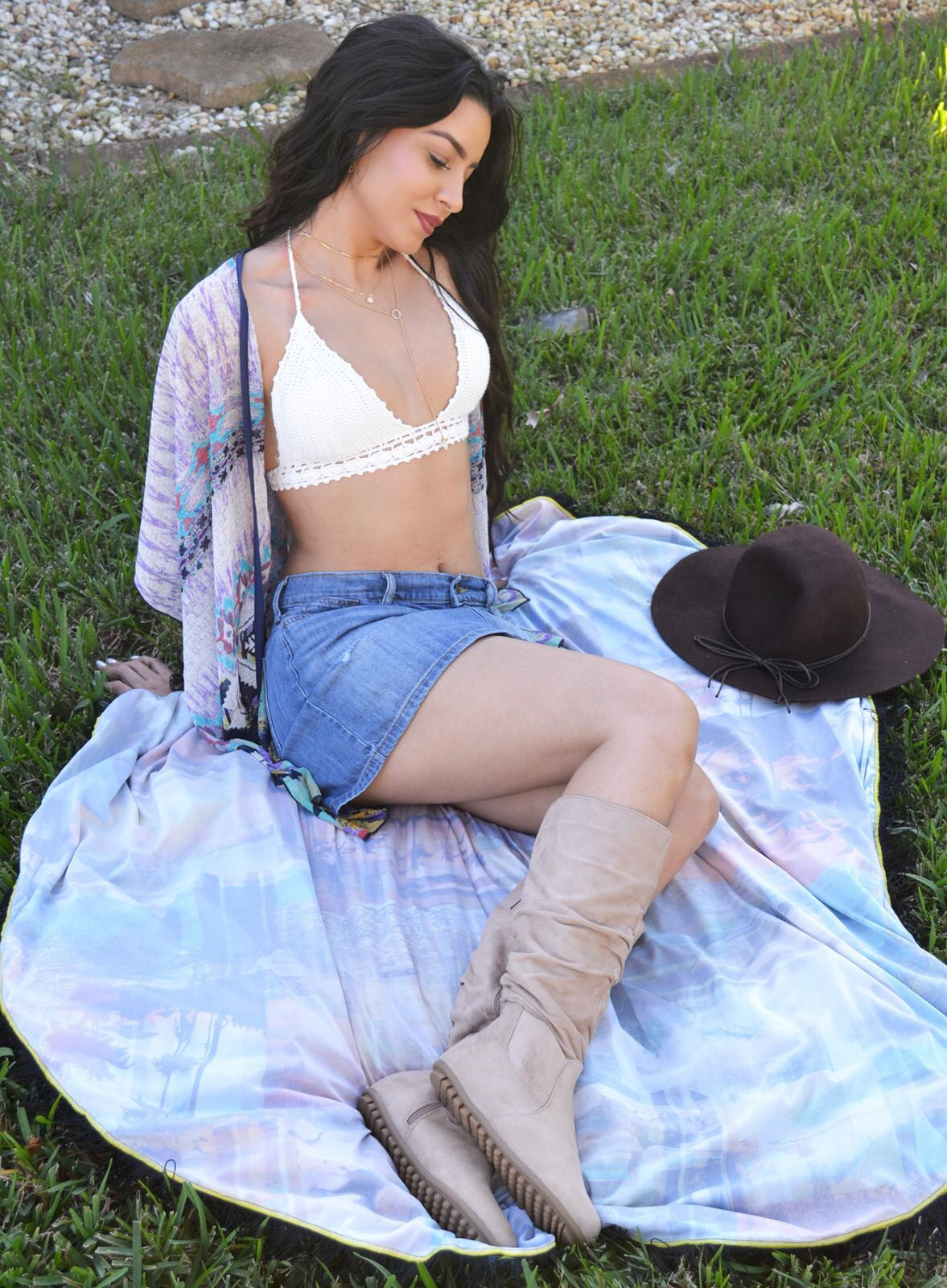 Woman sitting on grass wearing a white top, denim shorts, and beige boots with a hat nearby.