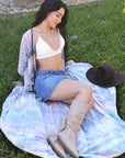 Woman sitting on grass wearing a white top, denim shorts, and beige boots with a hat nearby.