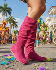 Person wearing pink calf-high boots in a festive street setting with colorful costumes and confetti.
